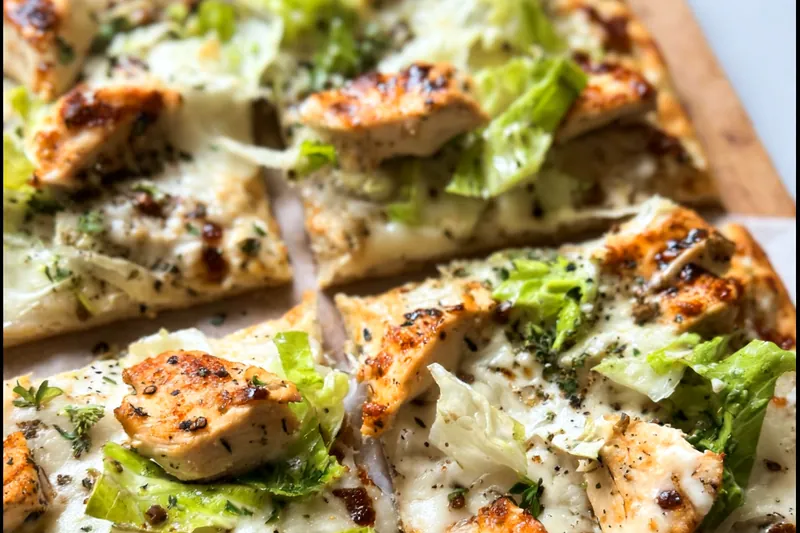 Chicken Caesar flatbread being assembled with grilled chicken and fresh romaine lettuce being added to the crispy flatbread base