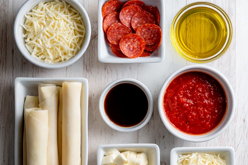 Ingredients for air fryer pizza rolls including pizza dough, sauce, shredded mozzarella, diced pepperoni, and fresh herbs arranged on a wooden cutting board
