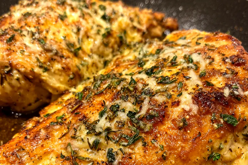 Herb and cheese crusted chicken breasts sizzling in a cast iron skillet with golden-brown crust developing, showing perfect searing technique